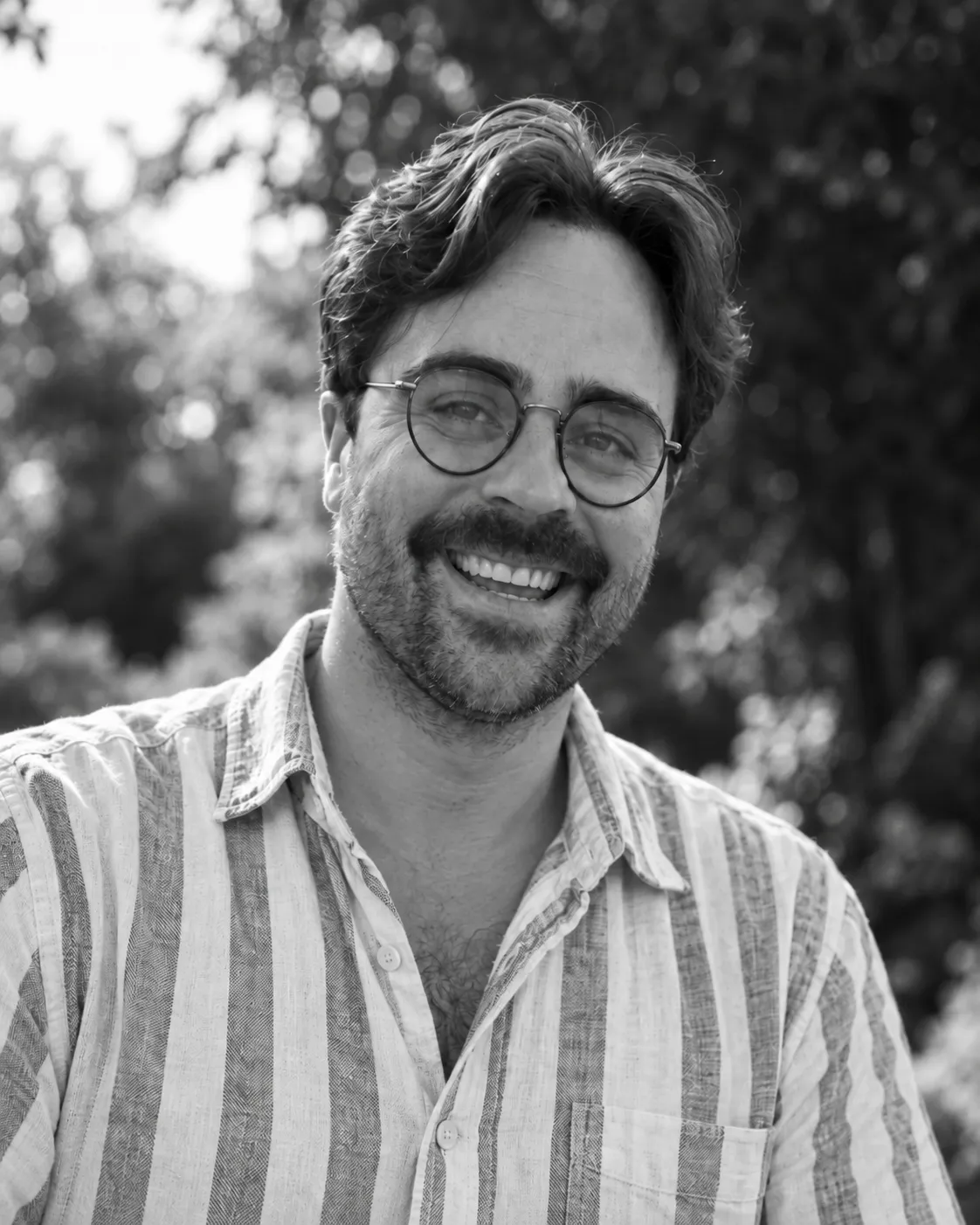 Joel, smiling, in round glasses and a striped shirt, with trees behind him.